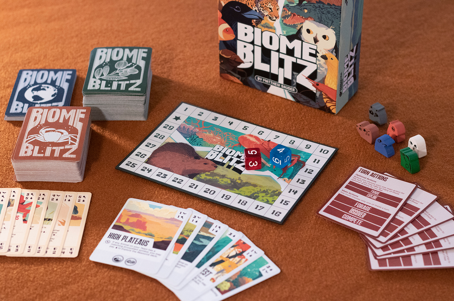 A photo of the Biome Blitz game box and contents. This includes a board, two dice, up to 80 animal cards, 18 ecosystem cards, 6 wooden animal meeples, and 6 turn action cards.